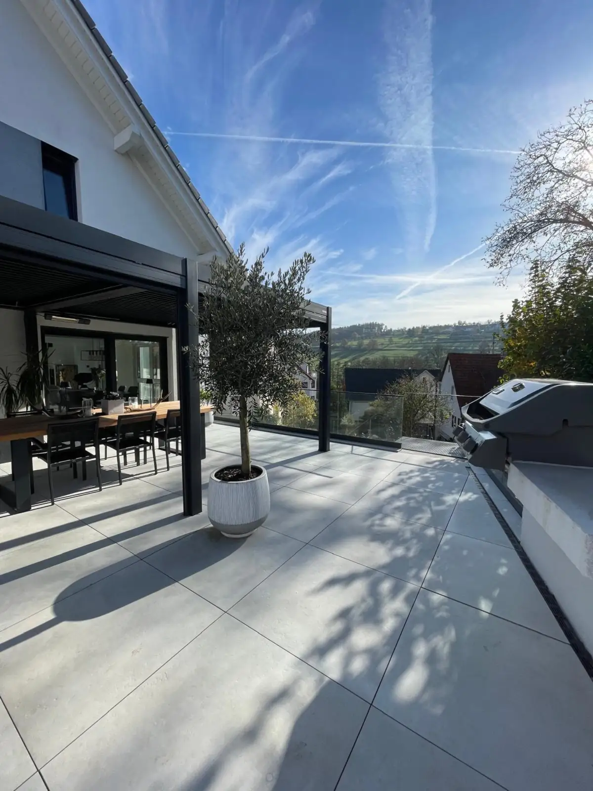 Terrasse aus großformatigen Platten mit Pergola, Esstisch und Olivenbaum, im Hintergrund Hügel und Wiesen
